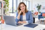 Frau mit Laptop und geschlossener Mappe an einem hellen Schreibtisch.